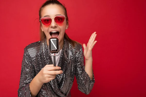 Zdjęcie piękne emocjonalnie pozytywne brunetka szczęśliwa dziewczynka noszenie stylowy połysk kurtka i czerwone okulary stojące izolowane nad czerwonym tle ściany śpiewając piosenkę do srebra — Zdjęcie stockowe