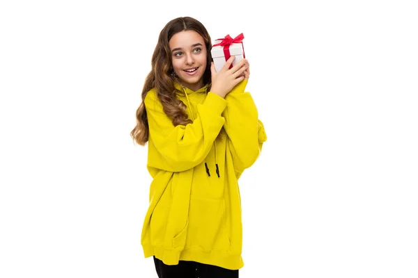 Foto de una hermosa adorable positiva feliz sonrisa morena adolescente en una elegante sudadera con capucha deportiva amarilla sosteniendo un regalo en una caja blanca con una cinta roja aislada sobre un fondo blanco con gratis —  Fotos de Stock