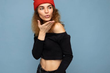 Genç, pozitif, hoş, esmer, kıvırcık saçlı kadının fotoğrafı. Samimi duyguları olan, şık, siyah bir bluz giyen ve mavi arka planda izole edilmiş kırmızı şapkalı bir kadın. Fotokopi alanı ile öpüşüyor.