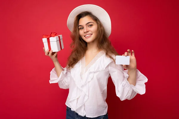 Портрет позитивной веселой модной женщины в формальной одежде с подарочной коробкой и кредиткой, смотрящей на камеру на красном фоне с копировальным пространством — стоковое фото