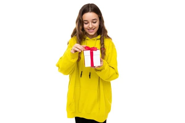 照片中，一个漂亮可爱的、积极的、快乐的、面带微笑的黑发少女穿着时髦的黄色运动服，手里拿着一件礼物，装在一个白色的盒子里，盒子里有一条红色的缎带，白色的背景是免费的 — 图库照片