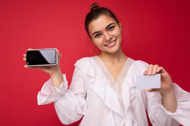 Resmî giysiler içinde pozitif neşeli bir kadının portresi ve cep telefonunu boş ekranla gösterme ve kırmızı arka planda izole edilmiş kameraya bakarak kesilen kredi kartı.