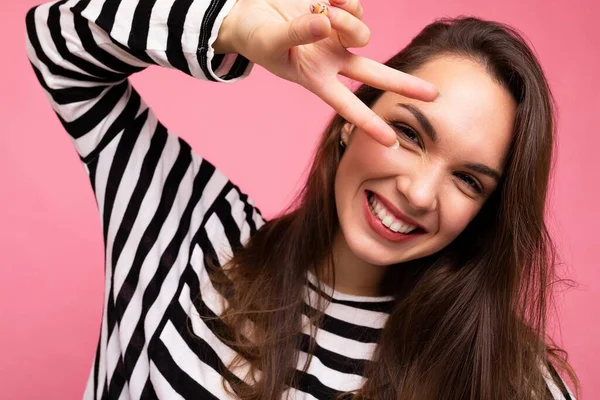 Foto de cerca de la joven europea positiva sonriente guapa feliz hermosa morena mujer con emociones sinceras usando jersey de rayas casuales y mostrando gesto de paz eith dos dedos aislados en —  Fotos de Stock