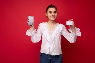 Beyaz bluz giyen, güzel Avrupalı, şık esmer bir kadın ve kırmızı arka planda izole edilmiş, samimi duygularla izole edilmiş şık bir şapka. Basit ve doğal bir şekilde kameraya bakıyor. Tutuluyor