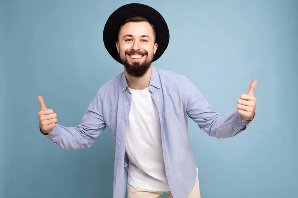 Kuva laukaus positiivinen onnellinen hymyilevä hyvännäköinen nuori ruskeaverikkö parrakas mies yllään rento sininen paita ja valkoinen t-paita ja tyylikäs musta hattu valmis eristetty sinisellä pohjalla tyhjä tila — kuvapankkivalokuva