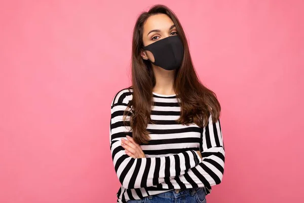 Zdjęcie młodej pięknej brunetki w masce ochronnej wirusa wielokrotnego użytku na twarzy przed koronawirusem izolowanym na różowej ścianie tła z wolną przestrzenią na tekst — Zdjęcie stockowe