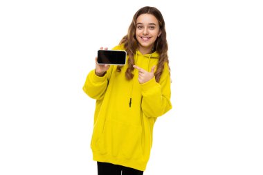 Güzel, sevimli, gülümseyen, neşeli, esmer, şık sarı spor kapüşonlu bir kız. Elinde siyah, boş bir arka planda izole edilmiş bir cep telefonu var.