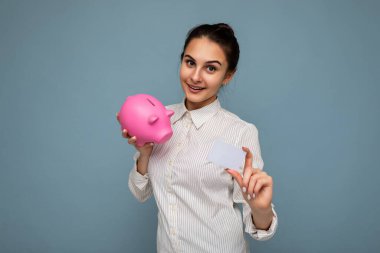 Mutlu, pozitif gülümseyen, samimi duyguları olan hoş, esmer bir kadının portresi. Fotokopi alanı olan mavi dar bir alanda izole edilmiş şık bir gömlek giyiyor. Para ve kredi için pembe domuz kutusu tutuyor.