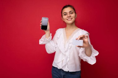 Beyaz bluz giyen, güzel Avrupalı, şık esmer bir kadın ve kırmızı arka planda izole edilmiş, samimi duygularla izole edilmiş şık bir şapka. Basit ve doğal bir şekilde kameraya bakıyor. Tutuluyor