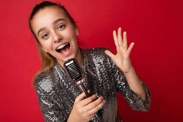 Shot van mooie emotionele positieve brunette kleine meisje dragen stijlvolle glans jurk geïsoleerd over rode achtergrond muur zingen van het lied in de zilveren microfoon op zoek naar camera — Stockfoto