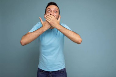Yetişkin, yakışıklı, sakalsız, sakalsız, samimi duyguları olan, mavi tişörtlü bir adamın fotoğrafı. Mavi arka plan duvarına izole edilmiş, boş bir alan ve elleriyle ağzını kapatan bir adam.
