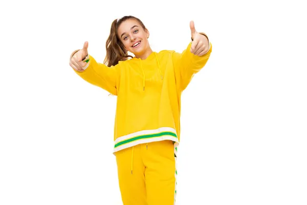 Zdjęcie pięknej uśmiechniętej uroczej dziewczyny z ciemnymi włosami z kucykiem w stylowo żółtym dresie z zielonymi paskami pokazuje palce do kamery odizolowanej na białym tle z wolną przestrzenią — Zdjęcie stockowe