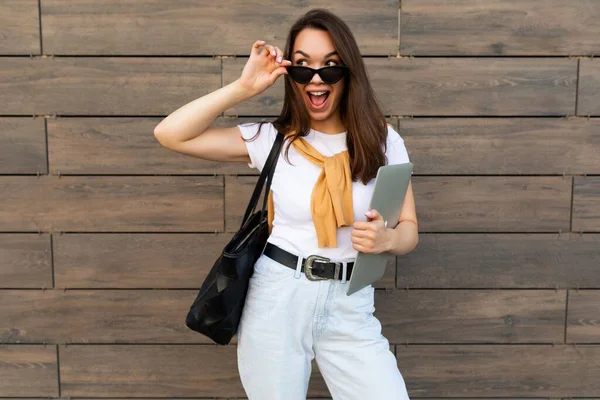 Photo of fascinating beautiful smiling happy positive charming young brunette girl wearing stylish clothes holding computer laptop and black sunglasses in white t-shirt and light blue jeans in the — Stock Photo, Image