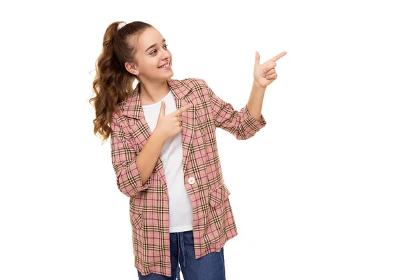 Photo de belle mignonne adorable fille adolescente brune souriante avec queue de cheval dans une veste à carreaux élégante et un jean bleu isolé sur fond blanc montre les doigts sur l'espace libre pour le texte — Photo