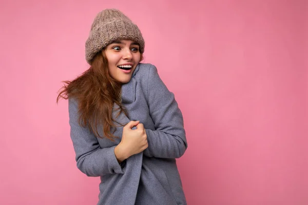 Fotografie krásné mladé šťastné brunetky žena izolované přes růžové pozadí zdi na sobě šedý podzimní kabát a šedý teplý klobouk při pohledu do strany a pocit chladu — Stock fotografie