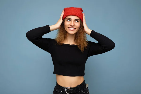 Φωτογραφία του νεαρού θετική χαμογελώντας χαριτωμένο όμορφη μελαχρινή γυναίκα σγουρά με ειλικρινή συναισθήματα φορώντας κομψό μαύρο καλλιέργεια κορυφή και κόκκινο καπέλο απομονώνονται σε μπλε φόντο με αντίγραφο χώρο — Φωτογραφία Αρχείου