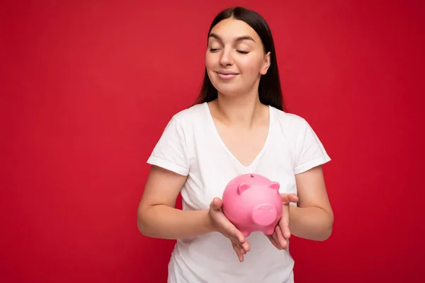 Shot of satisfied positive cute young beautiful attractive brunette woman with sincere emotions wearing casual white t-shirt isolated over the red wall with empty space and holding pink pig savings box — Stock fotografie