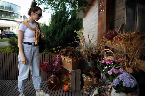 Photo of european beautiful attractive brunette woman wearing stylish white t-shirt and blue jeans chooses flowers as a gift in a street garden stall in the town — Stock Photo, Image