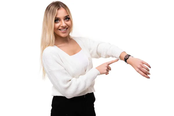 Foto de uma bela mulher loira sorrindo positivo muito surpreendente feliz jovem com maquiagem em uma camisola de malha branca elegante e calças pretas aponta um dedo para o relógio em sua mão isolado em um — Fotografia de Stock