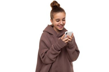 Güzel, pozitif saçlı, kahverengi kapüşonlu, kapüşonlu, elinde cep telefonu olan beyaz bir arka planda izole edilmiş sosyal ağlarda mesajlaşan güzel bir kızın fotoğrafı.
