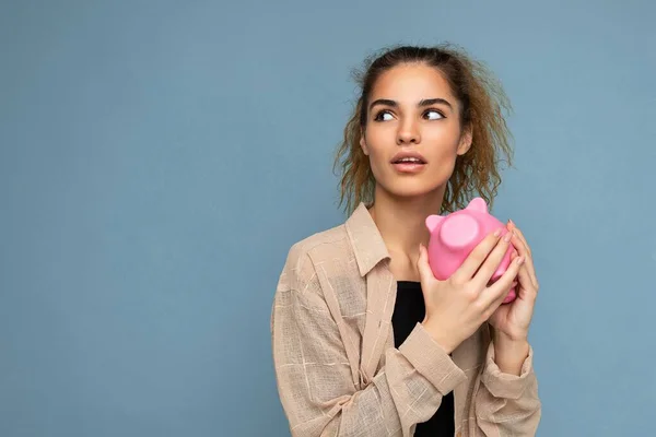 Foto von niedlichen jungen schönen attraktiven Frau mit dunklem lockigem Haar in einem Pferdeschwanz trägt lässig beige Hemd isoliert über blauem Hintergrund mit Kopierraum und hält rosa Sparschwein Box für Münzen — Stockfoto