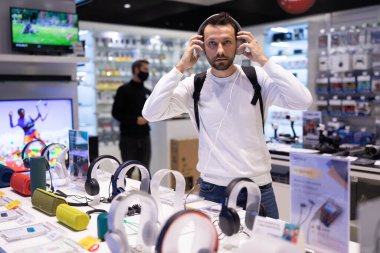 Minsk, Belarus - 27 Kasım 2021: Beyaz kazaklı ve sakallı yakışıklı bir esmer adamın fotoğrafı bir alışveriş merkezindeki elektronik mağazasından kulaklık seçiyor. Dükkan ve müzik konsepti