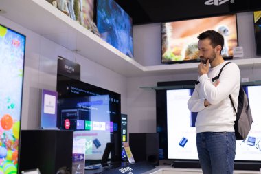 Minsk, Belarus - 27 Kasım 2021: Beyaz kazaklı, yetişkin, yakışıklı bir esmer adamın fotoğrafı bir alışveriş merkezindeki elektronik mağazasından televizyon seçiyor. Dükkan konsepti
