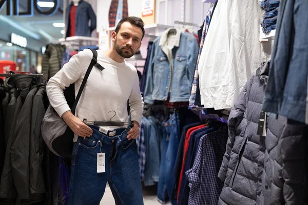 Adulto bonito morena homem em camisola branca escolhe jeans em uma loja de roupas em um shopping — Fotografia de Stock
