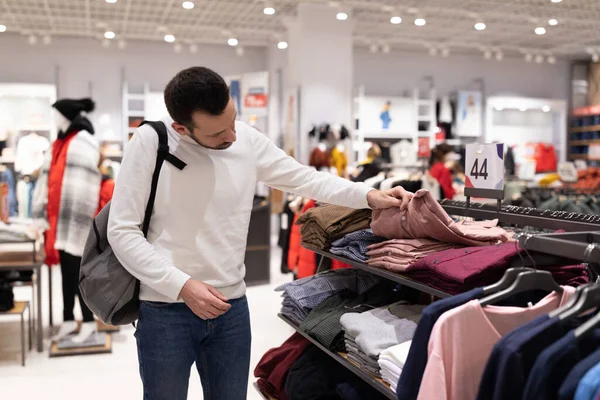 흰색 스웨터 를 입고 잘 생긴 성인 브루 우트 남자가 쇼핑 가게에서 유행하는 유행하는 셔츠 를 고르고 있다. 가게의 개념 — 스톡 사진