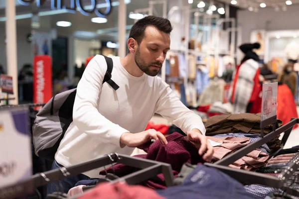Homem morena bonito adulto em camisola branca escolhe camisa elegante na moda na loja de roupas no shopping. Conceito de loja — Fotografia de Stock