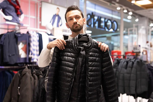 Ein gut aussehender, brünetter, unrasierter Mann wählt in einem Bekleidungsgeschäft in einem Einkaufszentrum eine warme schwarze Jacke. Oberbekleidung und Shop — Stockfoto