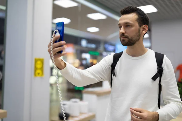 Foto de um homem morena bonito adulto com restolho em uma camisola branca escolhe um telefone celular azul em uma loja eletrônica em um shopping — Fotografia de Stock