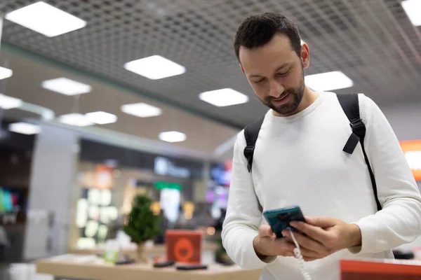 Foto de um adulto positivo bonito morena homem com restolho em uma camisola branca escolhe um telefone celular em uma loja eletrônica em um shopping — Fotografia de Stock