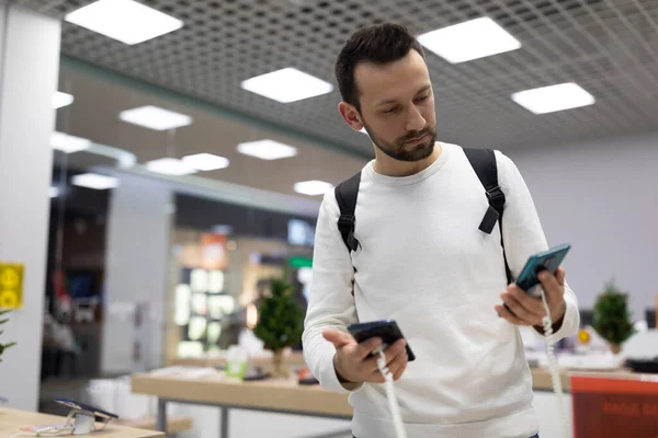 Foto de um homem morena bonito duvidoso adulto com restolho em uma camisola branca escolhe um telefone celular em uma loja eletrônica em um shopping — Fotografia de Stock