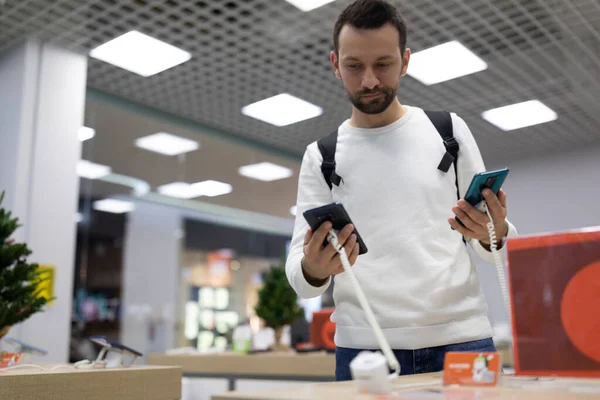 Foto de um adulto pensando bonito morena homem com restolho em uma camisola branca escolhe um telefone celular em uma loja eletrônica em um shopping — Fotografia de Stock