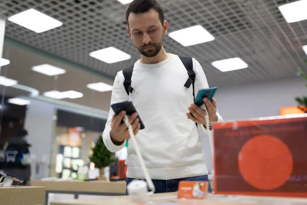 Foto de um homem morena pensativo bonito adulto com restolho em uma camisola branca escolhe um telefone celular em uma loja eletrônica em um shopping — Fotografia de Stock