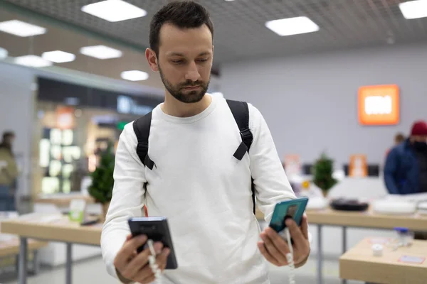 Foto de um homem morena bonito adulto com restolho em uma camisola branca escolhe um telefone celular em uma loja eletrônica em um shopping center. Conceito de loja — Fotografia de Stock