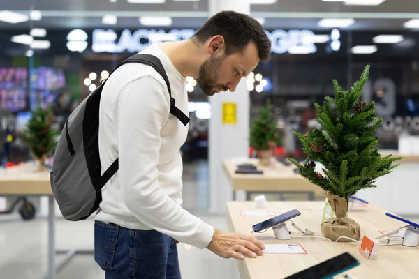 Foto de um homem morena pensamento duvidoso bonito adulto com restolho em uma camisola branca escolhe um telefone celular em uma loja eletrônica em um shopping center. Conceito de loja — Fotografia de Stock