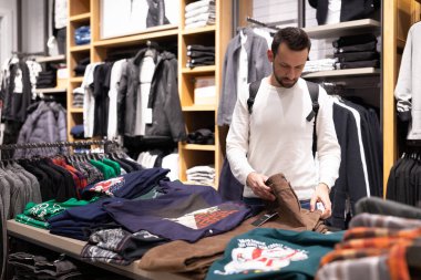 Minsk, Belarus - 27 Kasım 2021: Beyaz kazaklı, esmer bir adam alışveriş merkezindeki bir giyim mağazasından şık ve şık pantolon ve kot pantolon seçiyor. Dükkan