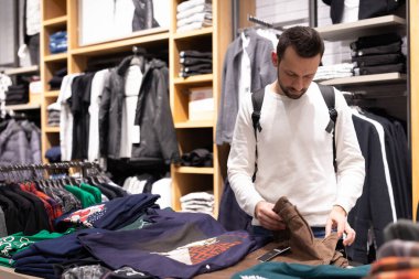 Minsk, Belarus - 27 Kasım 2021: Beyaz kazaklı, esmer bir adam alışveriş merkezindeki bir giyim mağazasından şık ve şık pantolon ve kot pantolon seçiyor. Dükkan