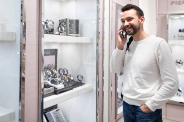 Minsk, Belarus - 27 Kasım 2021: Yetişkin, mutlu, mutlu, gülümseyen, beyaz kazaklı ve sırt çantalı bir esmer adamın fotoğrafı, alışveriş merkezindeki bir mağazadan şık bir kol saati seçiyor.