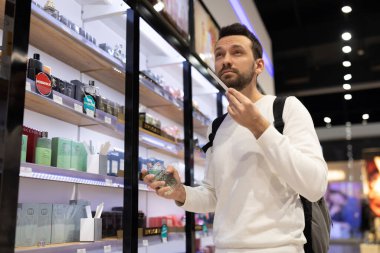 Minsk, Belarus - 27 Kasım 2021: Beyaz kazaklı, esmer, yetişkin, yakışıklı bir adamın fotoğrafı parfüm ve parfüm dükkanını bir alışveriş merkezinde seçiyor.