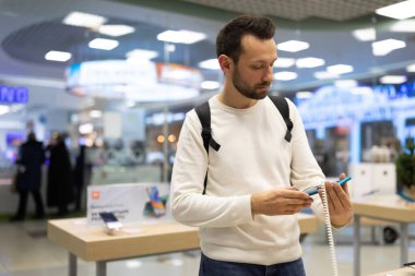 Minsk, Belarus - 27 Kasım 2021: Beyaz kazaklı, esmer, yetişkin bir adamın fotoğrafı bir alışveriş merkezindeki elektronik mağazasından cep telefonu seçiyor. Dükkan konsepti