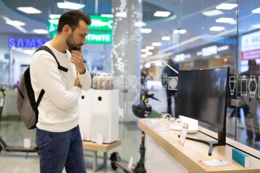 Minsk, Belarus - 27 Kasım 2021: Beyaz kazaklı ve sakallı yetişkin, yakışıklı bir esmer adamın fotoğrafı bir alışveriş merkezindeki elektronik mağazasından televizyon seçiyor. Dükkan konsepti