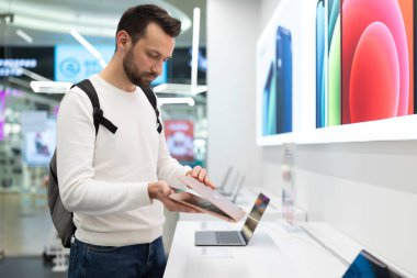 Minsk, Belarus - 27 Kasım 2021: Beyaz kazaklı, sakallı, yakışıklı, esmer bir erkeğin fotoğrafı iş için bir bilgisayar bilgisayarı seçiyor ve bir elektronik mağazasında okuyor.