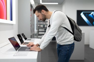 Minsk, Belarus - 27 Kasım 2021: Beyaz kazaklı, sakallı, yakışıklı, esmer bir adamın fotoğrafı iş için bir bilgisayar bilgisayarı seçiyor ve alışveriş merkezindeki bir elektronik mağazasında okuyor.