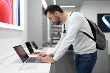 Minsk, Belarus - 27 Kasım 2021: Beyaz tişörtlü, sakallı, yetişkin, yakışıklı, kuşkulu, esmer bir adamın fotoğrafı iş için bir bilgisayar dizüstü bilgisayar seçiyor ve bir elektronik mağazasında çalışıyor.