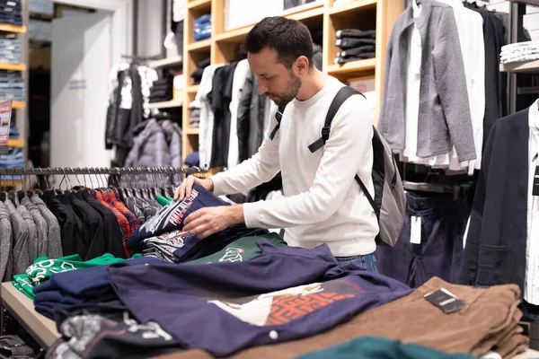 민스크, 벨로루시 - 노 브 27, 2021 년: 흰색 스웨트 셔츠를 입은 젊고 잘생긴 브루 넷 남자가 쇼핑 센터에서 유행하는 유행하는 옷을 선택 한다. 상점 — 스톡 사진