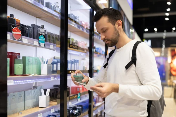 Minsk, Belarus - Nov 27, 2021: Foto de um homem morena bonito adulto com restolho em uma camisola branca escolhe perfumaria e adekolon em uma loja de perfume em um centro comercial — Fotografia de Stock