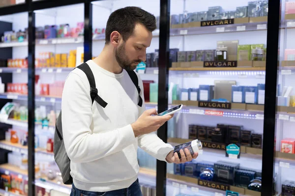 Minsk, Belarus - Nov 27, 2021: Foto de um jovem e duvidoso homem moreno bonito e atencioso com restolho em uma camisola branca escolhe perfumaria e adekolon em uma loja de perfumes em uma loja de compras — Fotografia de Stock